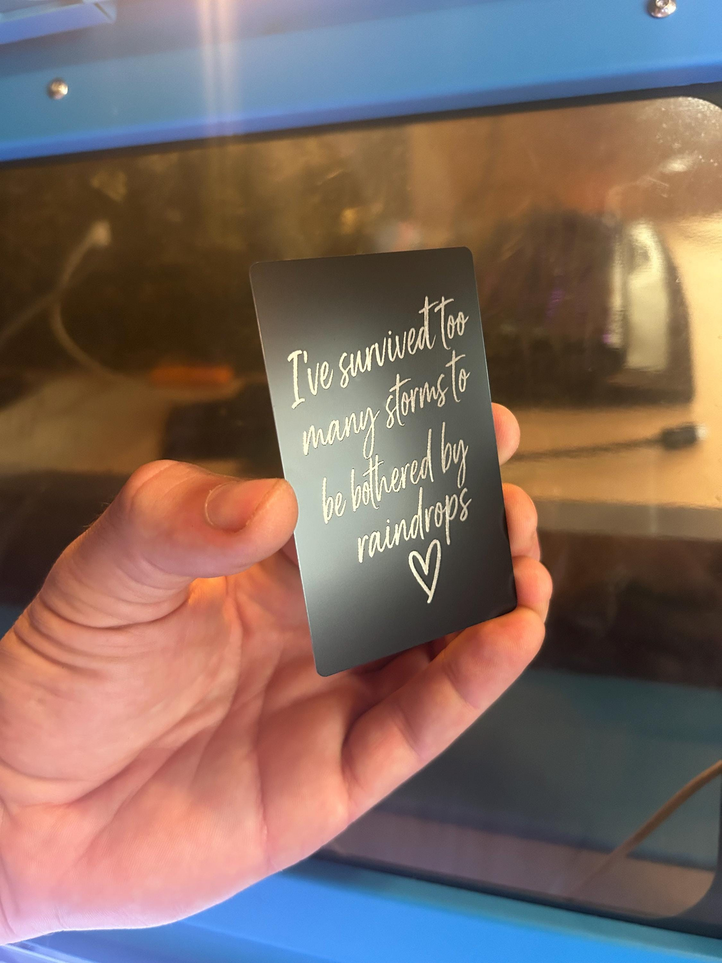 Person holding novelty laser cut metal quote card with “I’ve survived too many rainstorms…” engraving and heart graphic
