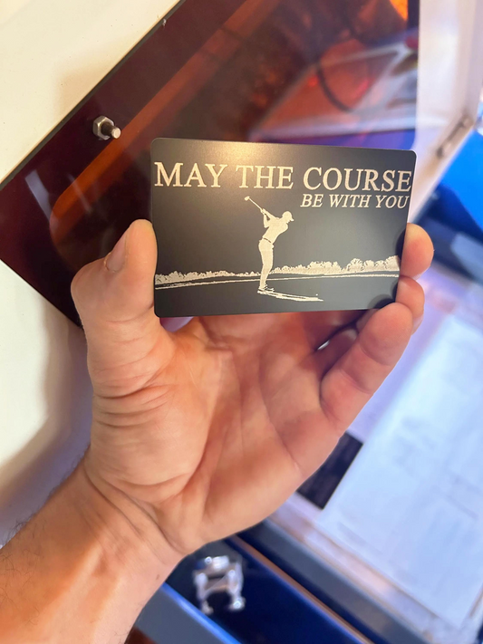 Hand holding a black/silver metal keepsake card with 'May the course be with you' text and golf graphic.