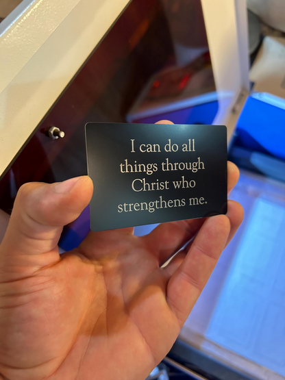 Hand holding a black card with "I Can Do ALL Things Through Jesus Christ" quote in front of a blurred background