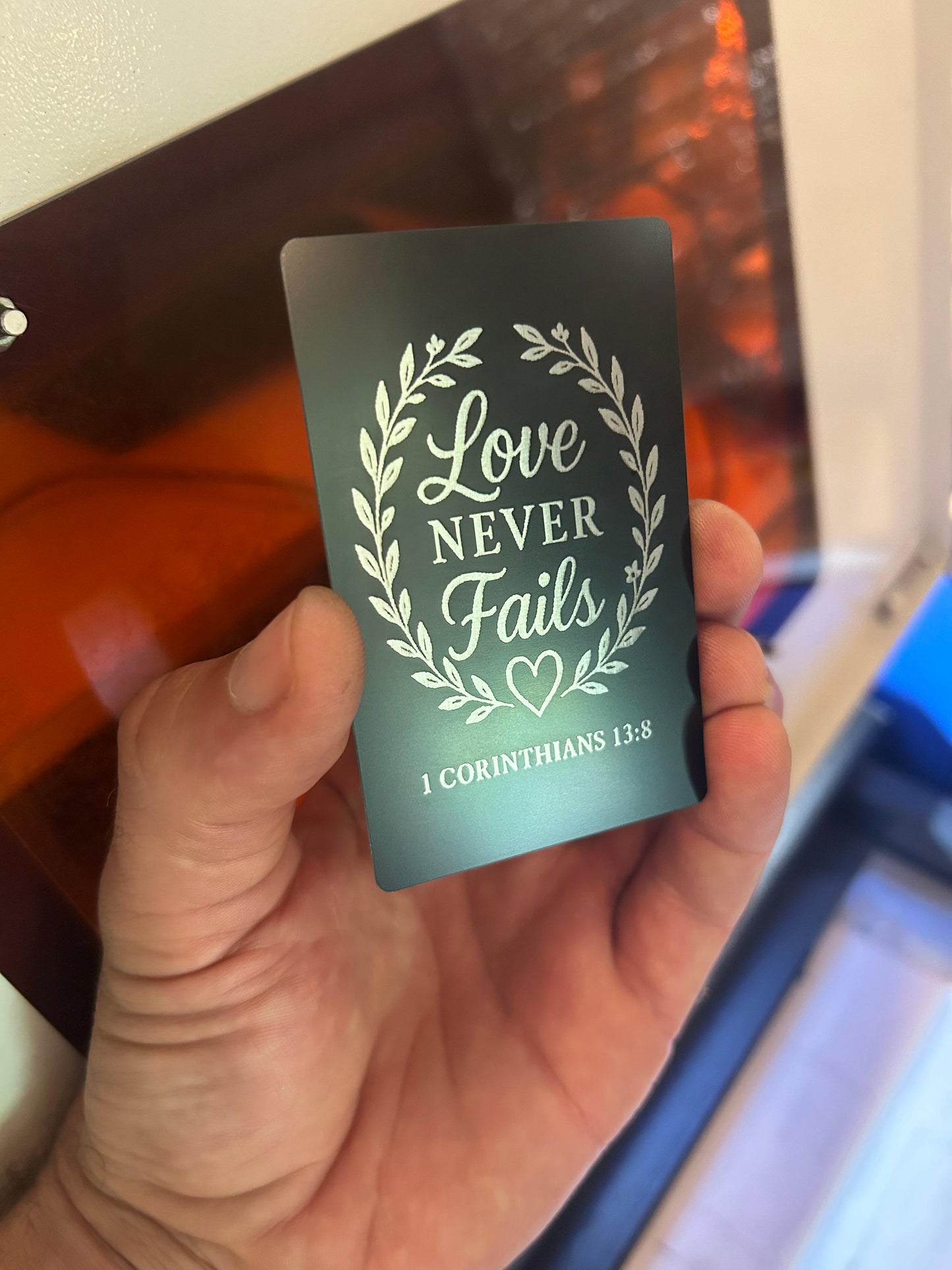 Laser engraved metal keepsake card with Love Never Fails heart artwork and 1 Corinthians 13:8 inscription below