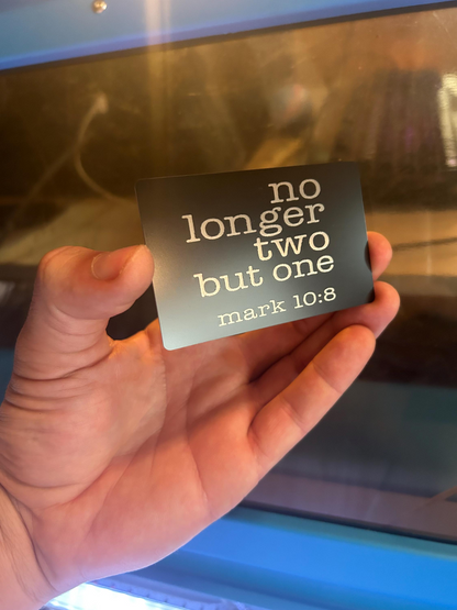Hand holding a black/silver metal wallet insert card with 'no longer two but one' and 'mark 10:8' text against a blurred background.