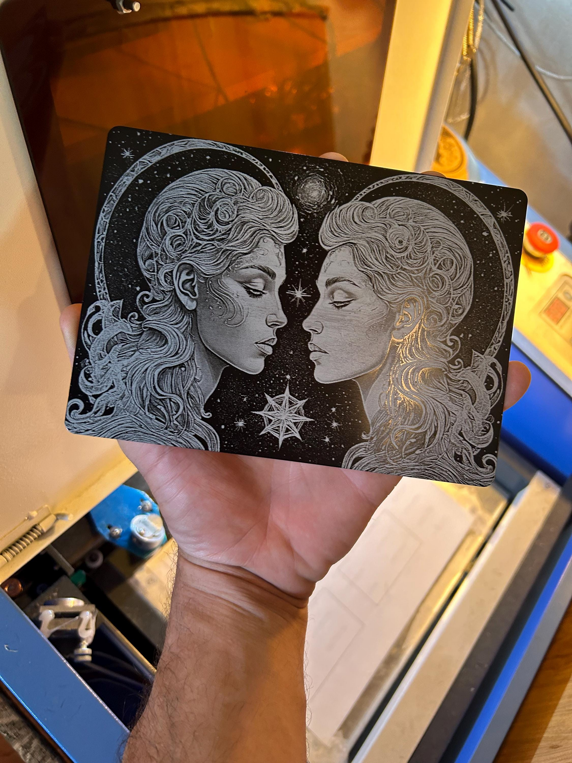 Intricate laser engraved design of two faces on a black metal plaque held by a hand with a blurred background.