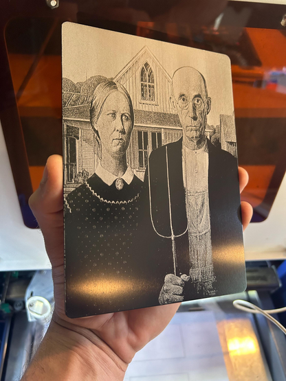 Metal card with engraved illustration of a couple and a house held in front of a blurred background.