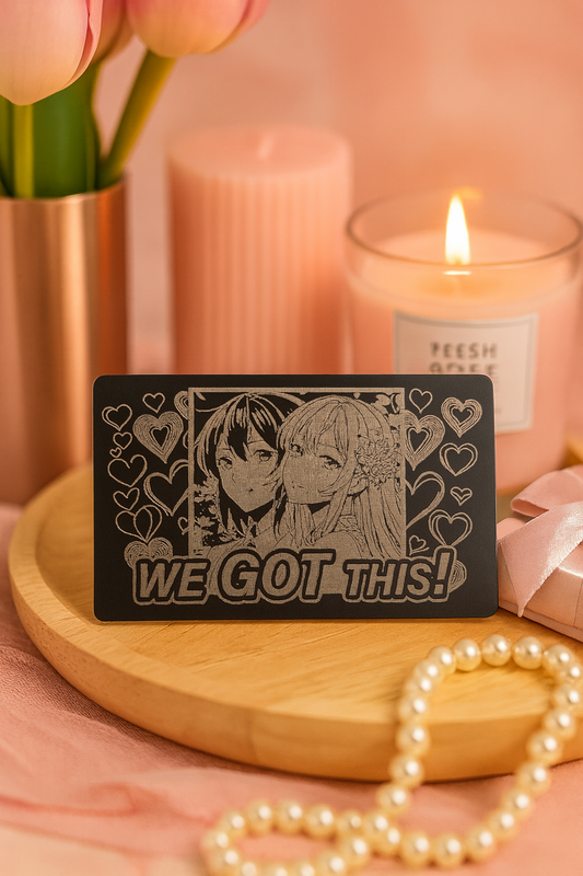 Metal laser engraved manga card with two anime characters and 'WE GOT THIS!' text on a wooden surface with candles and flowers in the background.