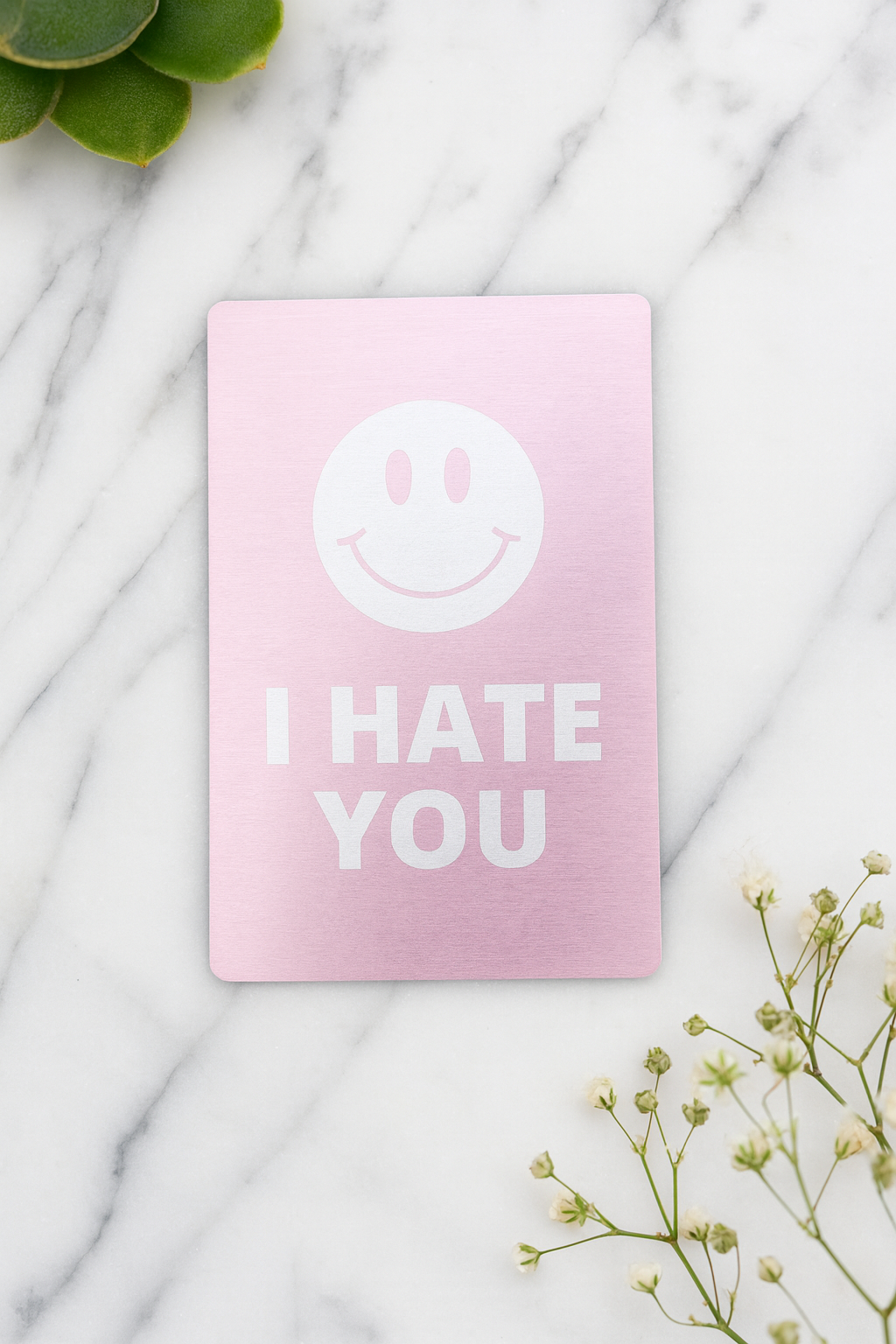 Rose gold metal keepsake card with a smiley face and 'I HATE YOU' text on a marble surface.