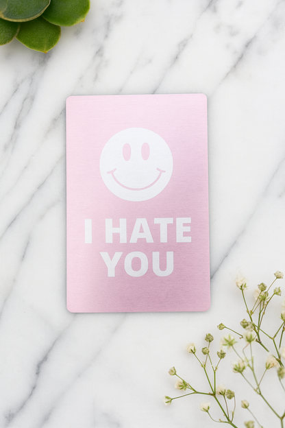 Rose gold metal keepsake card with a smiley face and 'I HATE YOU' text on a marble surface.
