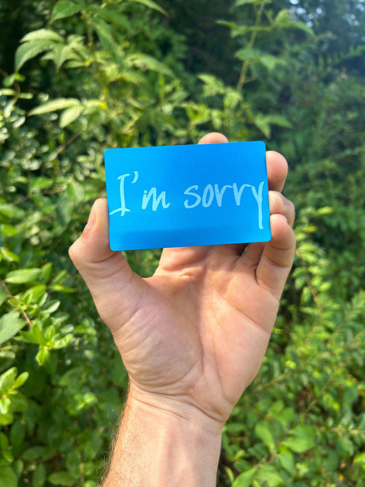 Hand holding a metal blue apology card with 'I'm sorry' text against a green leafy background