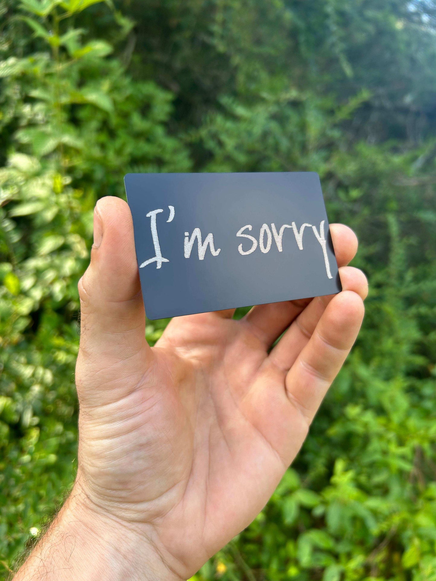 Hand holding a matte black/silver metal greeting card with 'I'm sorry' text against a green background