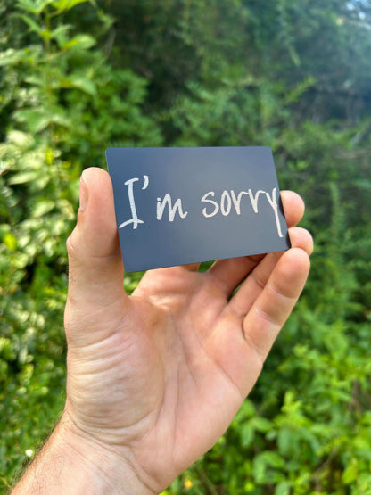 Hand holding a matte black/silver metal greeting card with 'I'm sorry' text against a green background