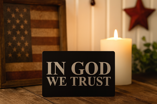 Decorative gift card with 'IN GOD WE TRUST' text laser engraved, photographed on a wooden surface with a candle and American flag in the background.