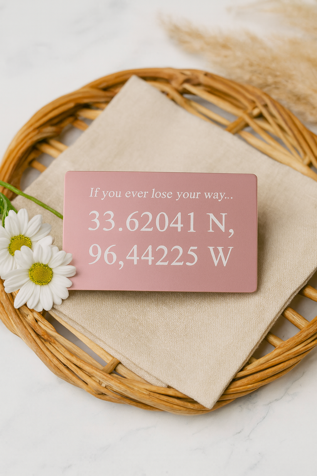 Pink card with custom laser engraved GPS coordinates on a beige napkin with flowers, placed on a woven mat.