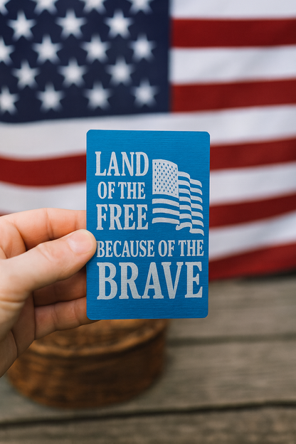 Blue metal keepsake card with 'Land of the Free Because of the Brave' text held in front of an American flag.