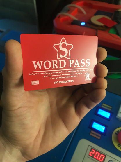Red/silver metal 'S WORD PASS' card held by a hand with a colorful laser engraving machine device in the background.