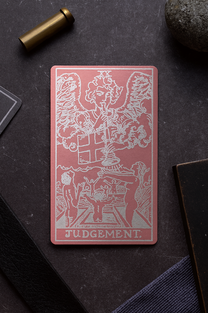 Pink metal tarot card titled 'Judgment' on a dark surface with a bullet and rock.