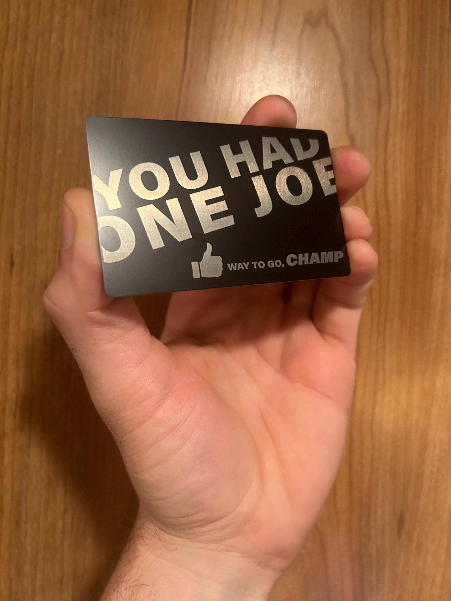 Hand holding a metal novelty gift card with 'You Had One Joe' text on a wooden surface