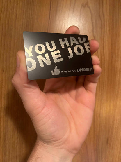 Hand holding a metal novelty gift card with 'You Had One Joe' text on a wooden surface