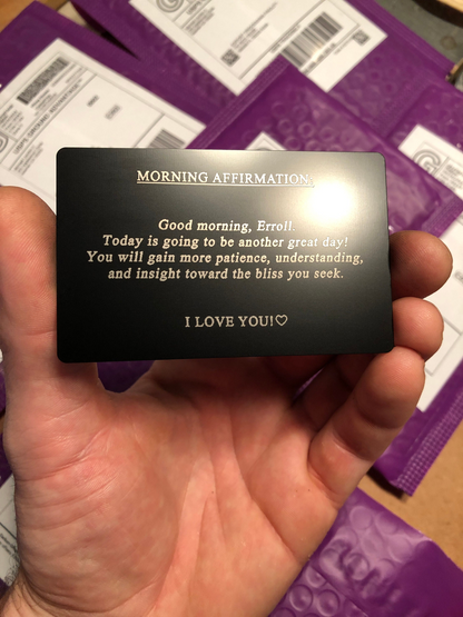 Hand holding a metal affirmations card with a personalized morning affirmation message example against a purple shipping department background.