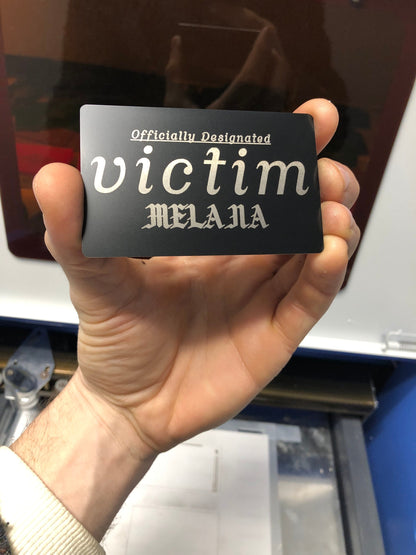 Metal gag gift card that reads "Officially designated VICTIM" with space for custom name engraving