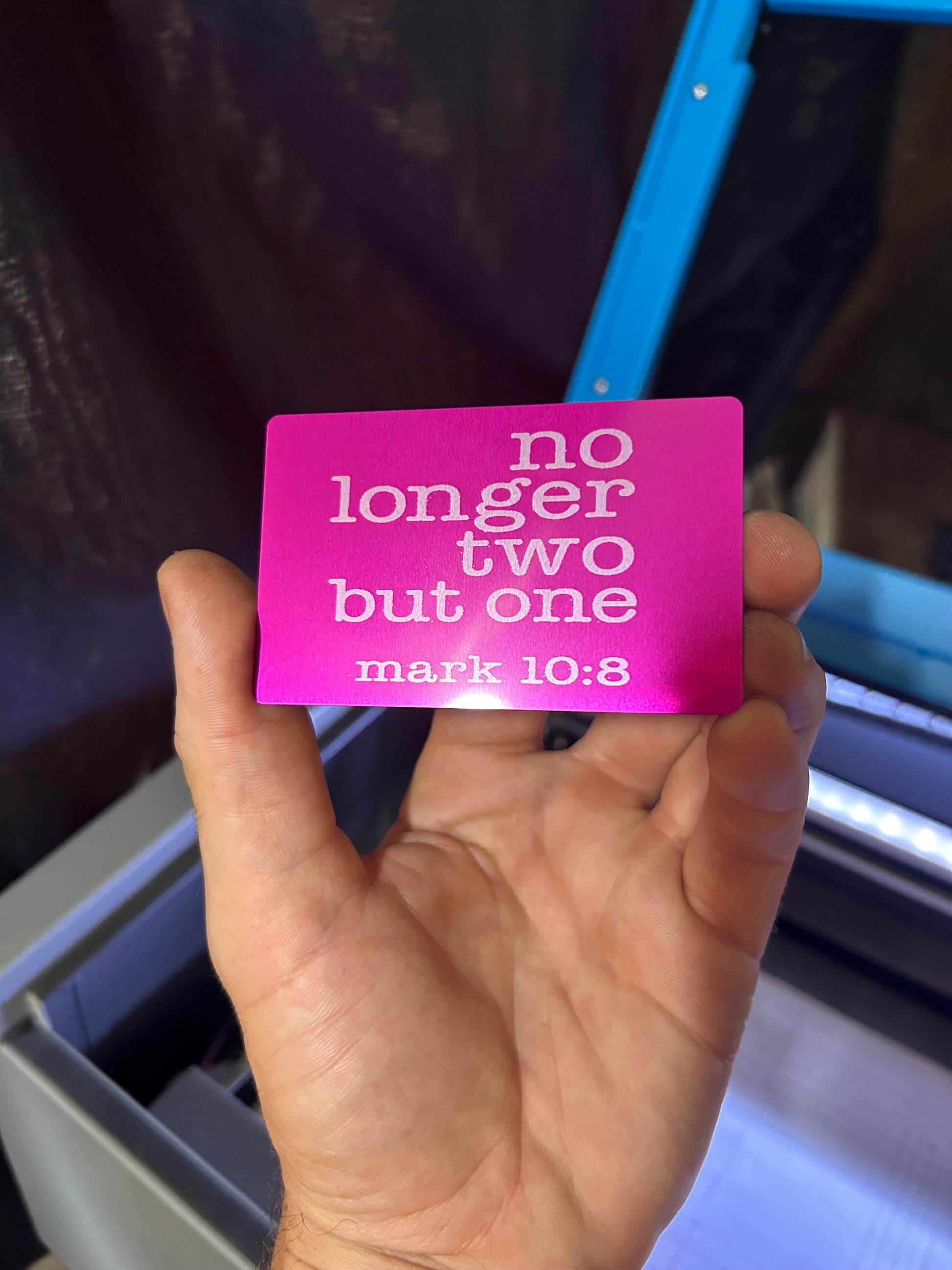 Hand holding a purple/silver metal gift card with text 'no longer two but one mark 10:8' against a blurred background