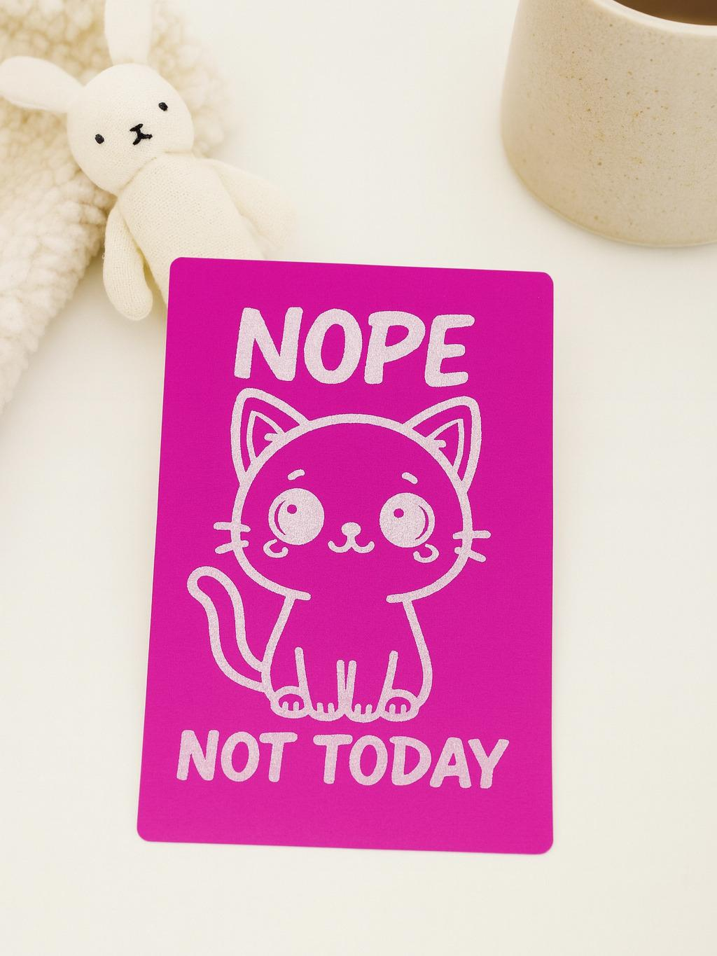 Pink card with a cat illustration and text 'NOPE NOT TODAY' on a white surface with a toy and mug.