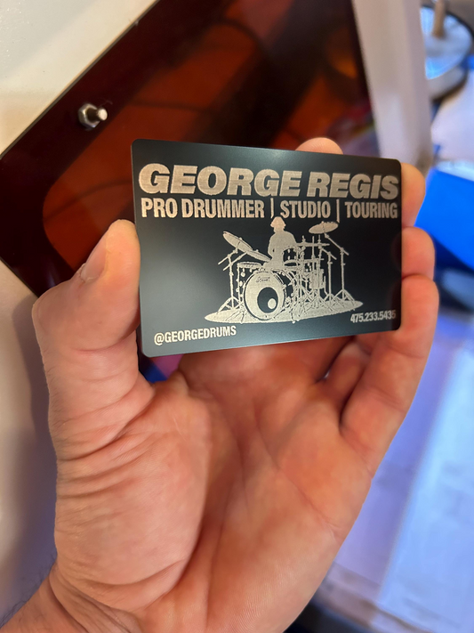 Hand holding a custom metal business card with 'George Regis Pro Drummer Studio Touring' text and drum set illustration.