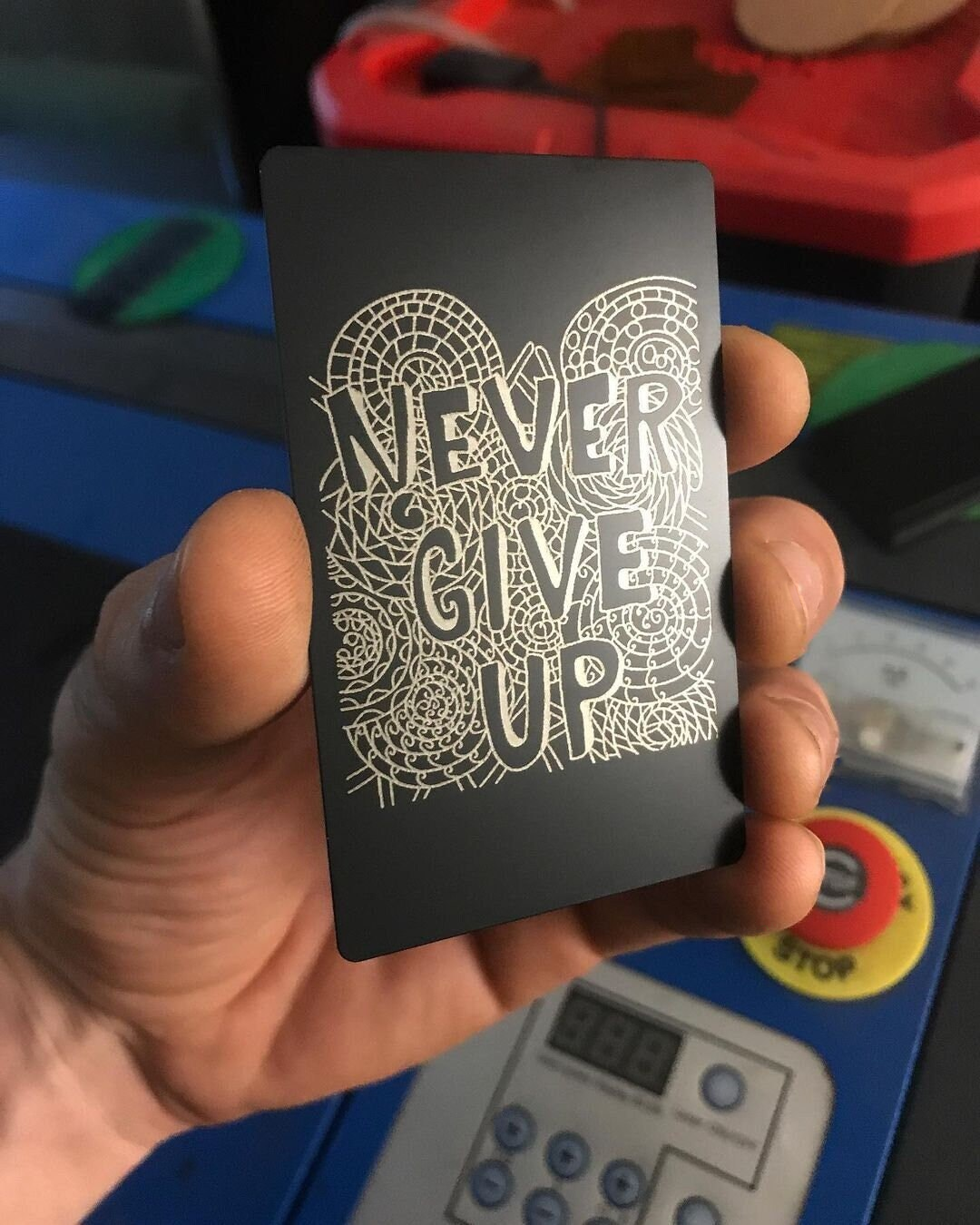 Hand holding a black metal keepsake card with 'Never Give Up' engraving against a colorful background