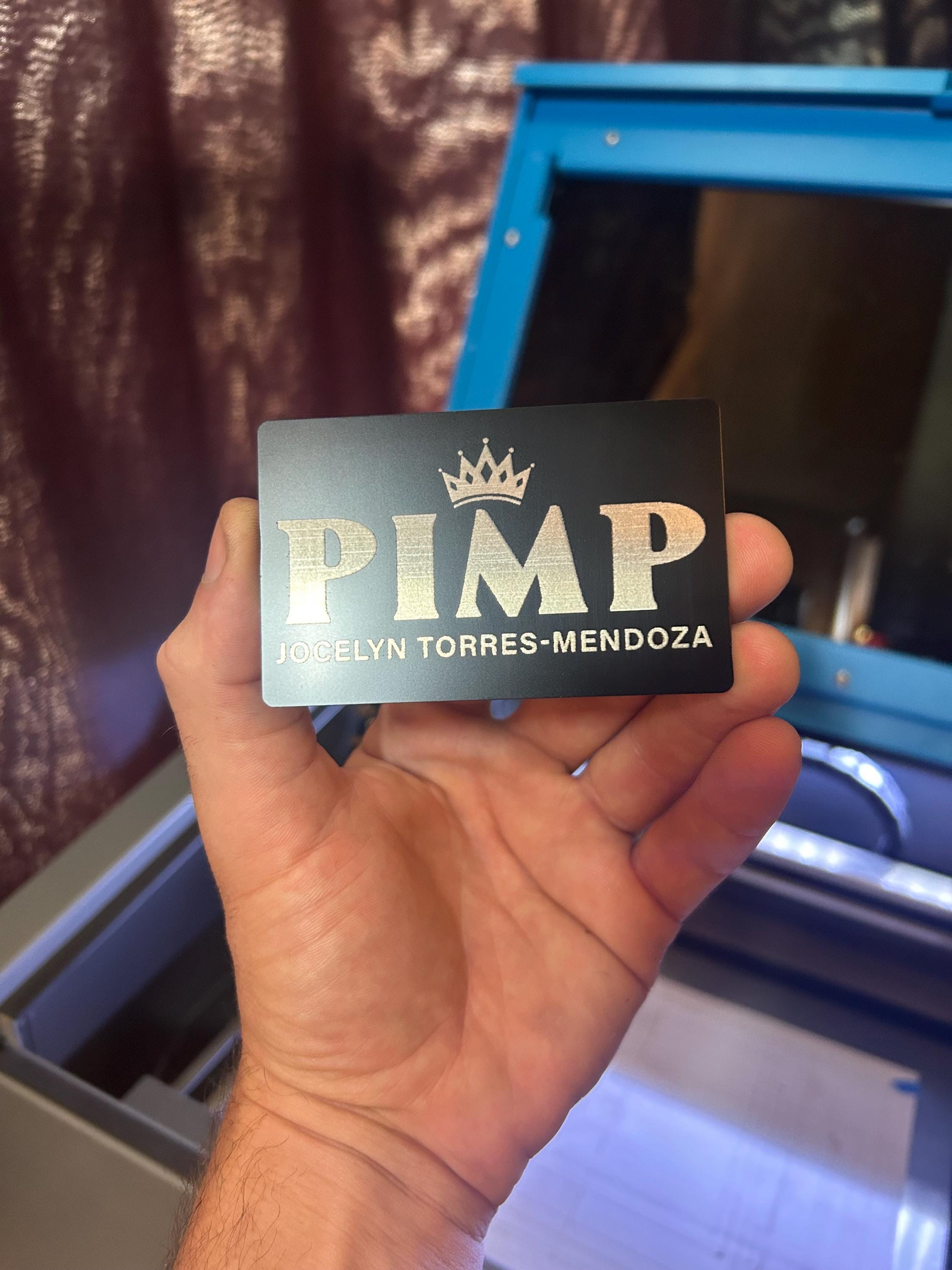 Hand holding a laser engraved metal keepsake gag gift card with 'PIMP' and a customized name printed on it, with a blurred background.