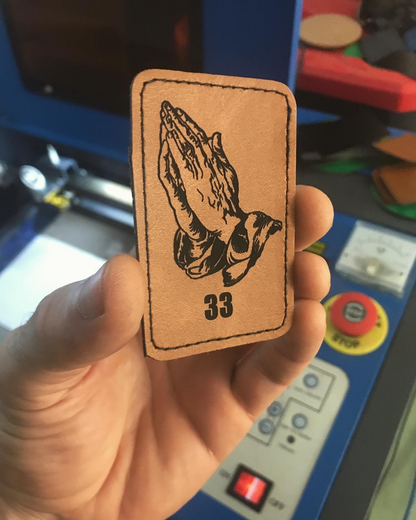 Brown leather patch with black praying hands design and 33 angel numbers held by a hand in front of a machine.