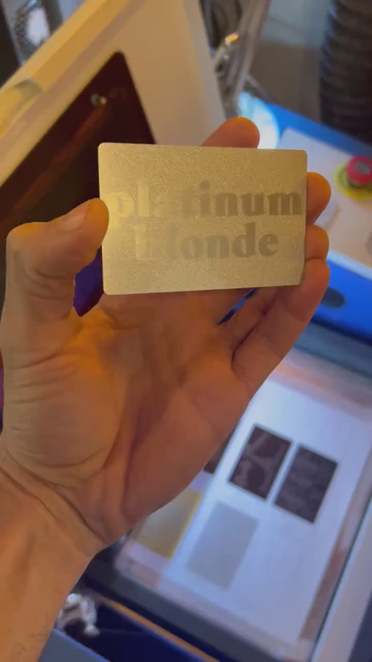 Engraved metal business card turning under light to highlight platinum blonde typographic design and reflective surface