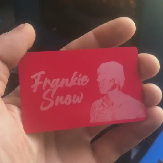 Hand holding a laser engraved red/silver metal business card with custom logo and text in front of laser engraving machines