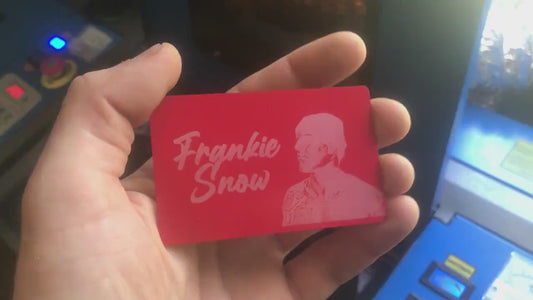Hand holding a laser engraved red/silver metal business card with custom logo and text in front of laser engraving machines