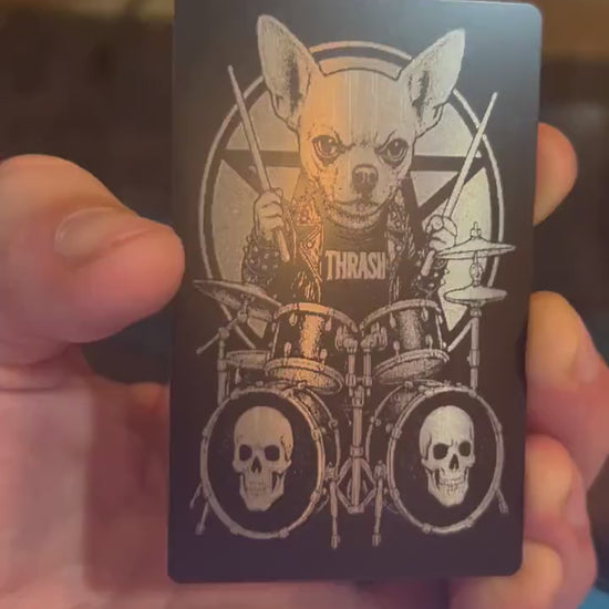 Hand holding a laser engraved black and silver funny keepsake card, with a cute dog heavy metal drummer design in front of a laser engraving machine