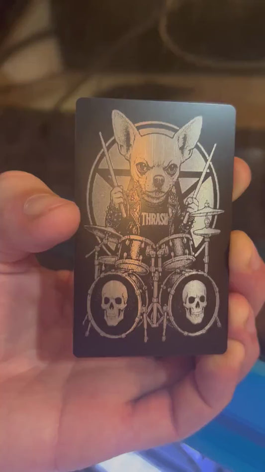 Hand holding a laser engraved black and silver funny keepsake card, with a cute dog heavy metal drummer design in front of a laser engraving machine