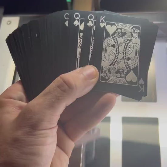 Hand holding a deck of 56 metal playing cards, with the King of hearts and 4 queens below, rotating in the light to demonstrate reflective qualities of metal in front of a laser engraving machine