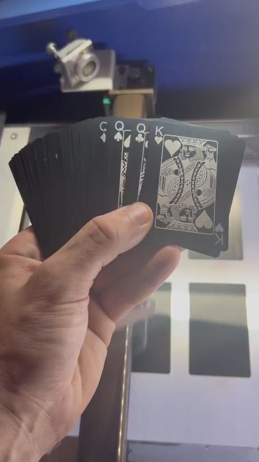 Hand holding a deck of 56 metal playing cards, with the King of hearts and 4 queens below, rotating in the light to demonstrate reflective qualities of metal in front of a laser engraving machine