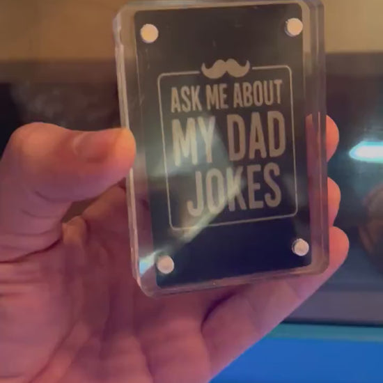 Hand holding a laser engraved meal gag gift card that reads "Ask Me About My Dad Jokes" with mustache design in black/silver anodized aluminum