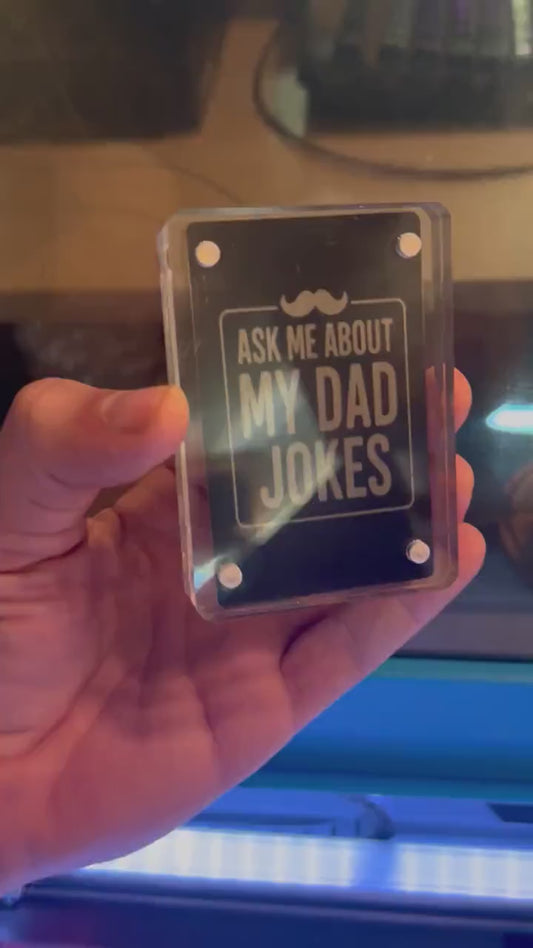 Hand holding a laser engraved meal gag gift card that reads "Ask Me About My Dad Jokes" with mustache design in black/silver anodized aluminum