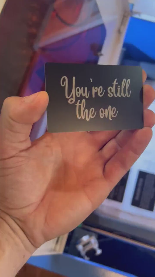 Video of a hand holding a romantic keepsake laser engraved gift card that reads: "You're Still The One", rotating in light to demonstrate reflective properties