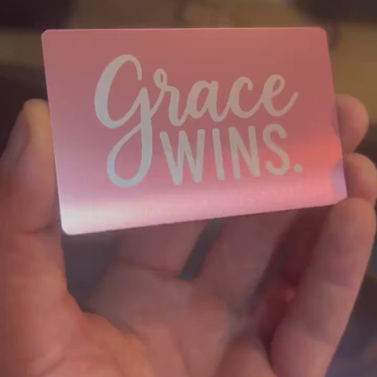 Hand holding a laser engraved metal keepsake card with "Grace Wins." text, rotating to show reflective qualities under light