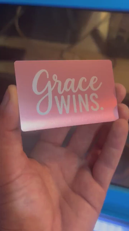 Hand holding a laser engraved metal keepsake card with "Grace Wins." text, rotating to show reflective qualities under light