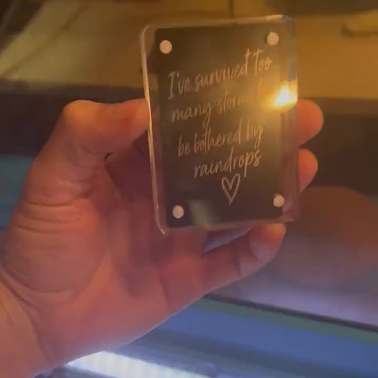 Hand showcasing a metal laser engraved keepsake card that reads "I've survived too many rainstorms to be bothered by a few rain drops", with a hand drawn heart graphic in black/silver anodized aluminum
