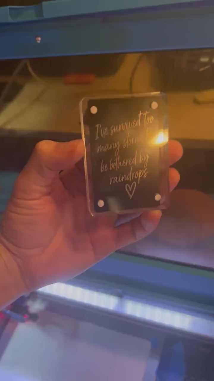 Hand showcasing a metal laser engraved keepsake card that reads "I've survived too many rainstorms to be bothered by a few rain drops", with a hand drawn heart graphic in black/silver anodized aluminum