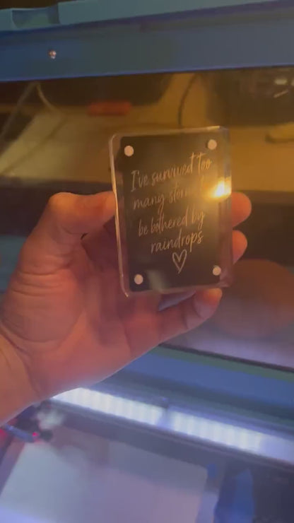 Hand showcasing a metal laser engraved keepsake card that reads "I've survived too many rainstorms to be bothered by a few rain drops", with a hand drawn heart graphic in black/silver anodized aluminum