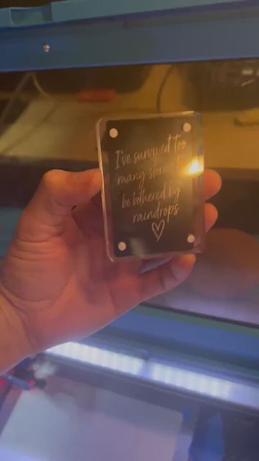 Hand showcasing a metal laser engraved keepsake card that reads "I've survived too many rainstorms to be bothered by a few rain drops", with a hand drawn heart graphic in black/silver anodized aluminum