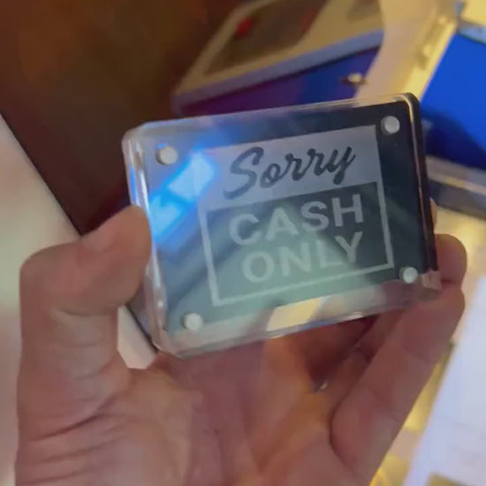 Hand holding a laser engraved black/silver Sorry Cash Only retail signage placard, shown in optional acrylic display case