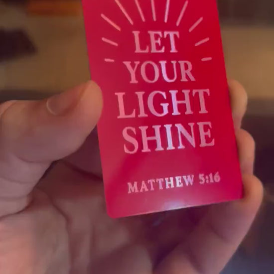 Close-up of hand showing engraved Bible verse card in red and silver metal with sun rays and Matthew 5:16 scripture reference