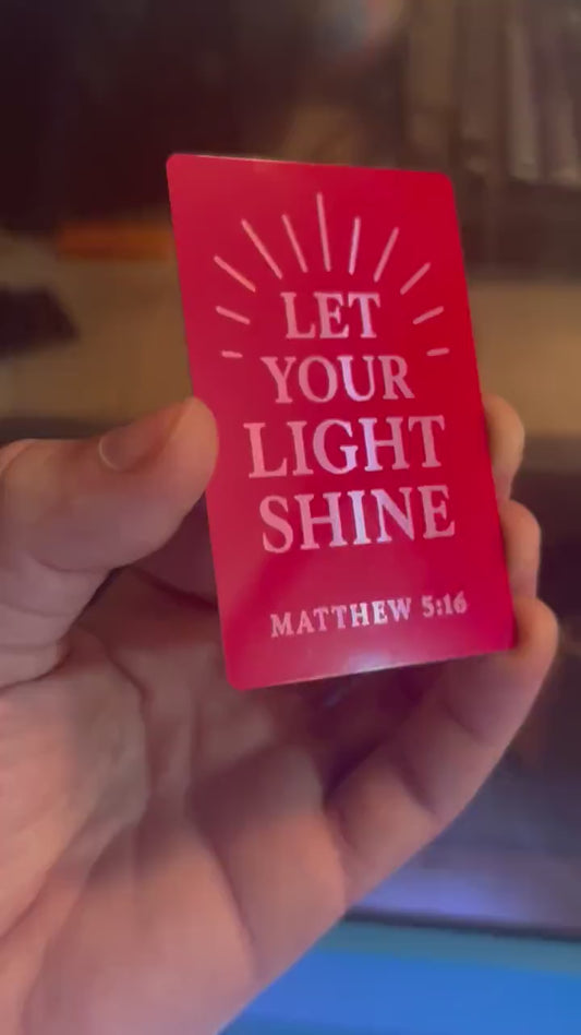 Close-up of hand showing engraved Bible verse card in red and silver metal with sun rays and Matthew 5:16 scripture reference