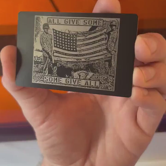 Hand holding a metal laser engraved anodized aluminum veteran keepsake, with patriotic "All Give Some, Some Give All" text and American flag soldier graphic
