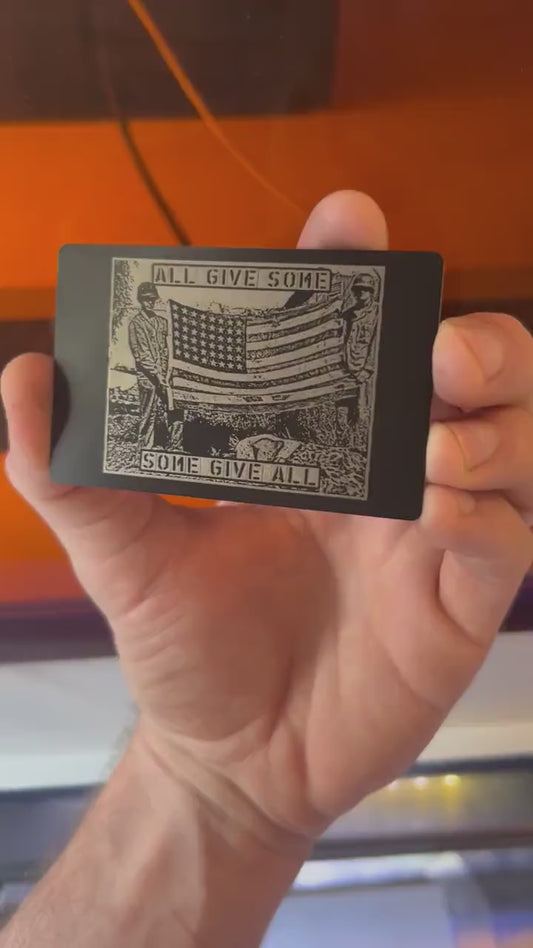 Hand holding a metal laser engraved anodized aluminum veteran keepsake, with patriotic "All Give Some, Some Give All" text and American flag soldier graphic