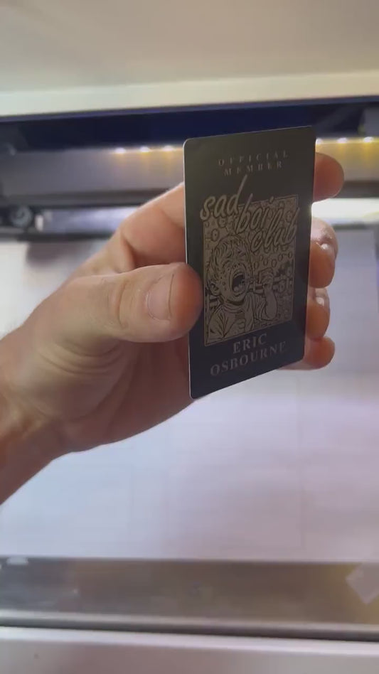 Hand holding laser engraved metal keepsake card with Sad Boi Club, a young boy blowing bubbles and crying, with personalized name engraving below